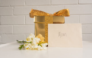 Elegant gold gift box for a Gold Cross necklace, with a 'Mom' card and delicate white flowers—perfect for Mother’s Day.