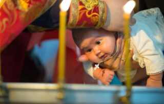Infant during Orthodox baptism wearing a Baptism gold cross necklace, held by priest with candles in the background.
